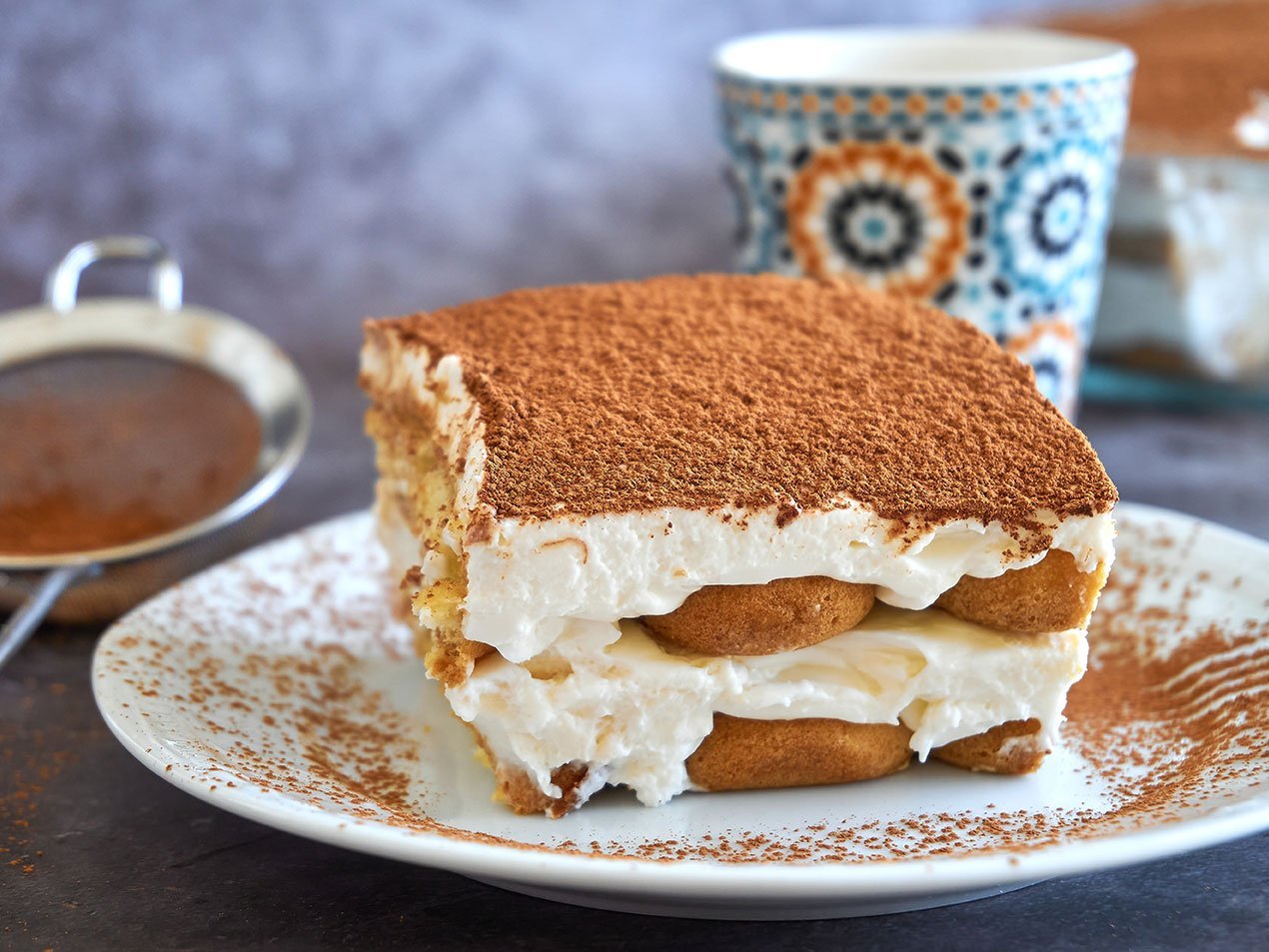 Square Slice Of Tiramisu Cake And A Cup Of Coffee In The Backgro