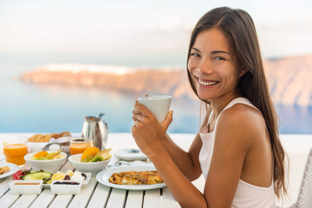 Breakfast Woman Eating Brunch At Luxury Travel Hotel Restaurant Drinking Coffee Cup Over Mediterranean View From Outside Balcony. Greek Healthy Food Santonini Vacation.