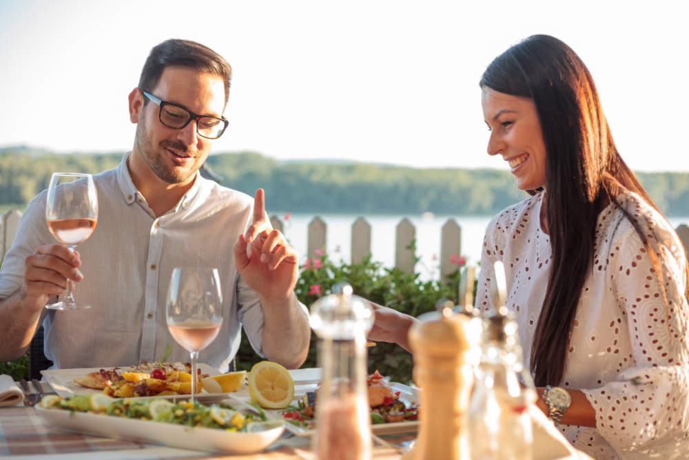 Happy Young Couple Eating Fish Dinner In Riverside Restaurant. Husband And Wife Celebrating Anniversary With Lunch In Restaurant. Having A Great Time, Talking And Laughing, Drinking Wine