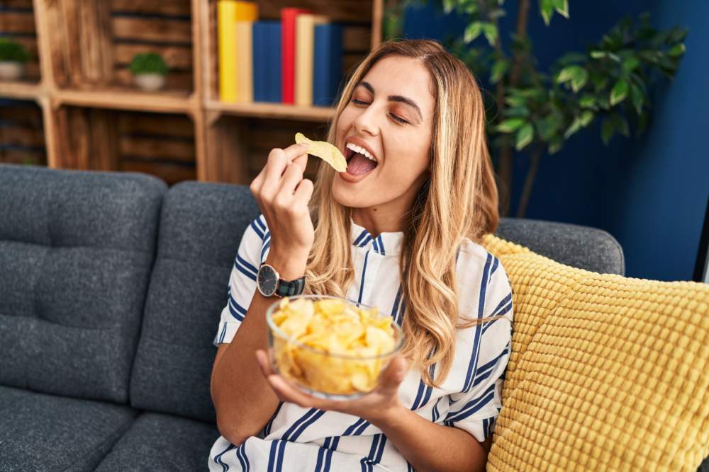 Young Blonde Woman Eating Chips Potatoes Sitting On Sofa At Home