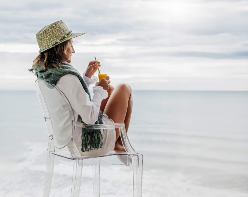 A Middle Aged Woman Sips An Orange Juice While Contemplating The Sea. Concept Of Well Being And Tranquility.