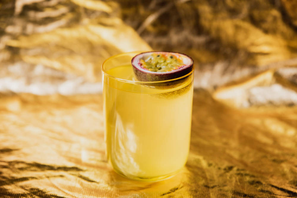 A Bright Yellow Cocktail With Ice In A Rocks Lowball Glass Garnished With Passion Fruit